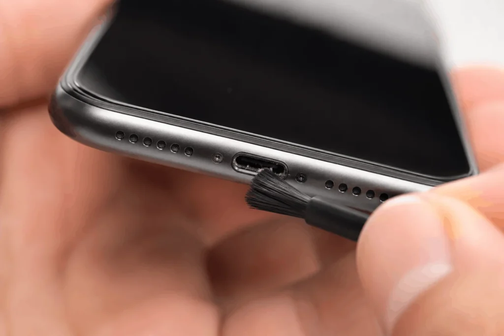 IPHONE Not Charging: Causes And Fixes That Work 3 Macro photo of a soft-bristled brush cleaning dust from an iPhone Lightning or USB-C charging port while the phone is powered off.